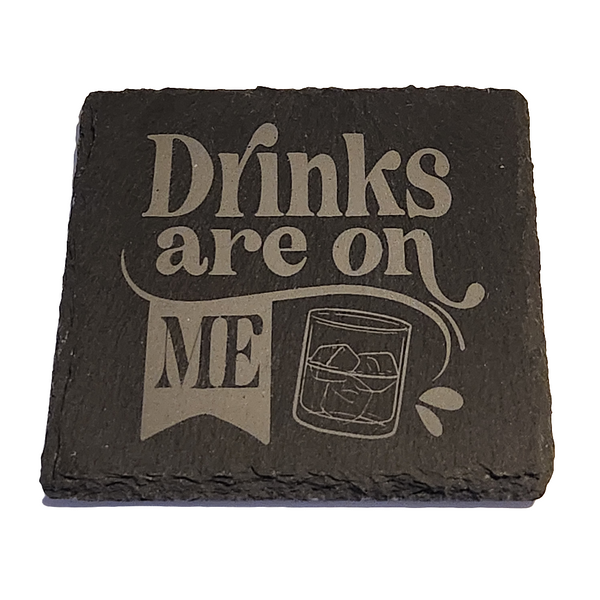 A black slate coaster with the phrase 'DRINKS ARE ON ME' engraved in large white letters, accompanied by an illustration of a glass with ice 