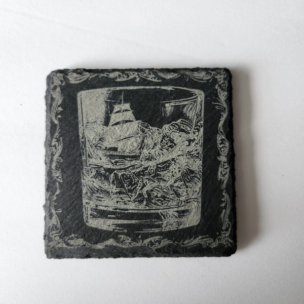 A black slate coaster with an engraved image of a ship in a glass.