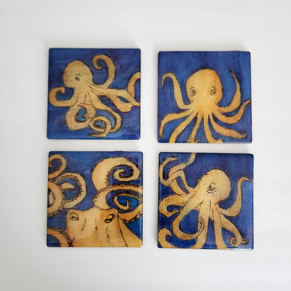 A set of four square coasters featuring hand-drawn and burned octopus designs on a blue background, with a cork back for each coaster.