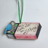 Hand painted wood ornament depicting a mouse next to a red and white gift box with text on it, with a green hanger.