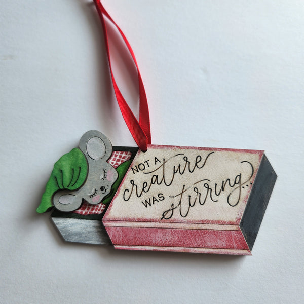 Hand painted mouse ornament on a red hanger, depicting a mouse inside a small open box with the text 'Not a creature was stirring.'.