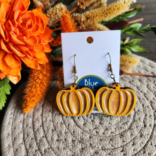 A pair of pumpkin-shaped earrings made from wood and resin, displayed on a card with 'Blue Bird' branding, surrounded by orange flowers.