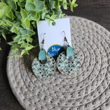 A pair of round floral earrings with leather accents and brass hooks, displayed on a textured surface, with a white card and 'Blue Bourbon Designs' branding in the background.