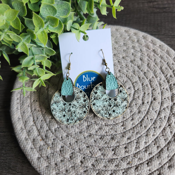 A pair of round floral earrings with leather accents and brass hooks, displayed on a textured surface, with a white card and 'Blue Bourbon Designs' branding in the background.
