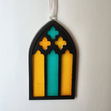 A resin stained glass window ornament with a black base and multicolored patterns, designed to look like stained glass on a ribbon hanger.