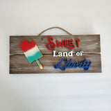 A novelty sign with a red and white popsicle on a wooden board with the text 'Sweet Land of Liberty' 