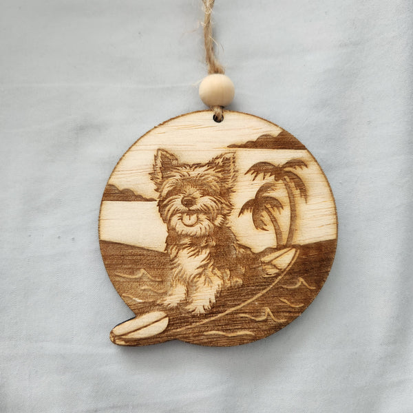 A circular wooden ornament featuring an engraved image of a Yorkie puppy on a surfboard, with a palm tree and sea in the background. It has a twine hanger and a wooden bead.